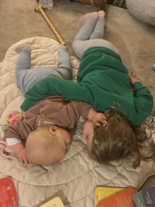 Two young girls — a baby and a toddler — lie on a blanket on the floor hugging each other. 