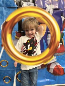 A 4-year-old boy stands behind a giant golden circle - as if the circle formed a picture frame. He's smiling broadly while looking through the circle, and has his left arm up and behind his head. 