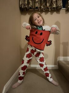 A young girl stands on what appears to be a stairwell. She wears white pajamas with red designs and appears to be holding up a red square with arms, eyes, and a smiling face. She's grinning for the camera.