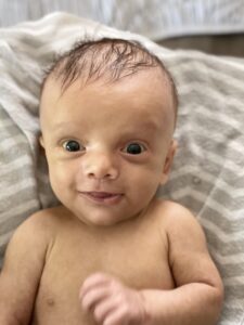 A baby boy lies on his back on a striped towel and smiles up at the camera.