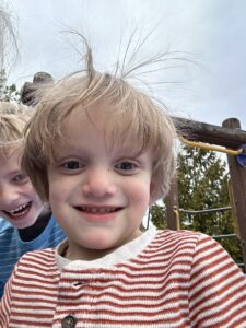 A young boy in a red-and-white-striped shirt smiles for the camera, with another young boy, his brother, standing behind him laughing. They're outdoors and the wind is blowing the first boy's hair straight up. 