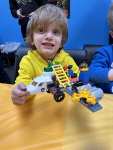 A 4-year-old boy with a full head of shaggy hair and a yellow hoodie shows off a Legos airplane he's made. 