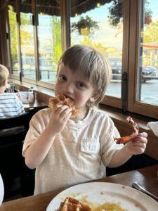 A young boy sits at a booth in a restaurant. He's taking a bite of a waffle that he's holding in his right hand, and he's holding a piece of bacon in the other hand. A plate in front of him has another piece of waffle covered in syrup. There are large windows to the boy's left, and it appears to be a sunny morning.