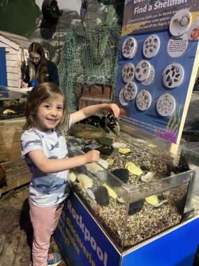 A young girl with brown hair stands and smiles before what appears to be an aquatic display.