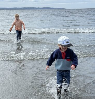 Two young boys run through gentle waves lapping on a beach. It looks to be a cloudy day, and both are wearing jeans and sweatshirts.