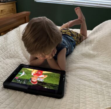 A young boy lies on his stomach on a white quilt. His hands are propping up his chin, and he's watching a video on a tablet in front of him.