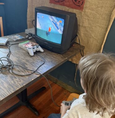 A blond-haired boy holds video game controls and appears to be playing an old-fashioned game on a small television set.