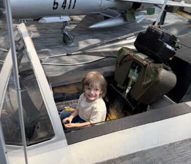 A young, blond-haired boy sits in what appears to be the cockpit of a stationary fighter jet. Another such jet is in the background.