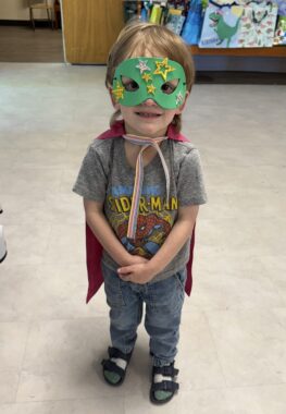 A blond-haired boy wears a green face mask with stars on it, as well as a red cape, a gray Spider-Man T-shirt, and blue jeans. He's standing indoors on what appears to be a light gray carpet.