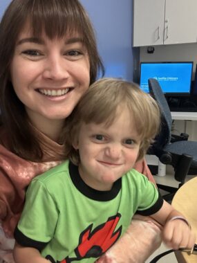 A young boy makes a funny face while sitting on his mom's lap in a clinical office. The mom is smiling.