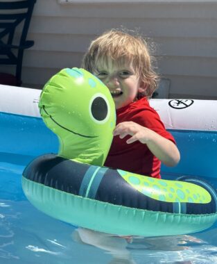 A young blond-haired boy in a red shirt plays in a small inflatable pool on his back deck. Around his waist is a small inflatable inner tube designed to look like a turtle.