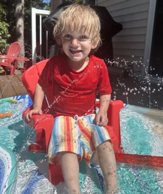 A young, blond-haired boy in a red shirt and striped swim trunks sits in a small red chair on top of a splash pad. There's water up to his ankles and several small jets shooting water up at him. He's smiling broadly at the camera.
