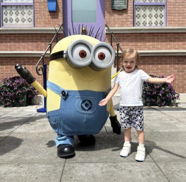 A 4-year-old boy poses for a photo with a Minion in front of a brick building at Universal Studios Hollywood. Both the boy and the character have their arms outstretched, and the boy is smiling broadly.