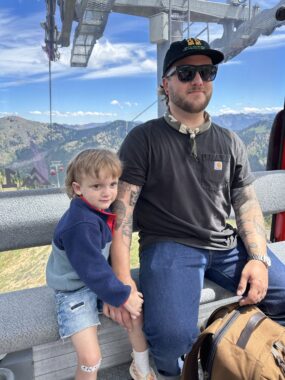 A young boy sits on a gondola ride and holds the hand of his young, bearded dad, who's wearing denim pants, a short-sleeve black shirt, a cap, and sunglasses. The boy, in denim shorts and a long-sleeve shirt, is leaning into his dad for protection, but has a coy smile on his face. It's a clear, sunny day, and majestic green mountains rise in the distance behind the two.