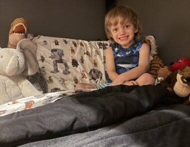 A boy smiles wide while sitting in bed. The sheets feature Lego Star Wars characters, and the boy, who wears a sleeveless shirt, is surrounded by stuffed animals and toys.