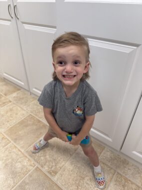 A 4-year-old boy smiles for a photo while standing with his legs spread wide. His blond hair has the shape of a mullet, and he's wearing a gray T-shirt, shorts, and rainbow checkered shoes. It looks like he's indoors, perhaps in his home, as he's standing on a tiled floor against white doors.