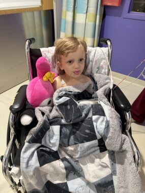 A blond 4-year-old boy sits in a wheelchair with a plush, checkered blanket covering him up to his chest.