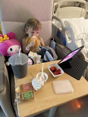 A 4-year-old boy sits in a large chair in a hospital room. On the table in front of him there's a yogurt container, some berries, and other food and drink items, as well as a tablet propped up so the boy can watch a show while he eats.