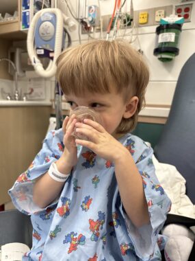 A 4-year-old boy holds a transparent mask to his face, covering his nose and mouth. He's in a light blue hospital gown with what appear to be cartoon characters on it, and is sitting in a chair in a medical setting. He looks to his right with smiling eyes.