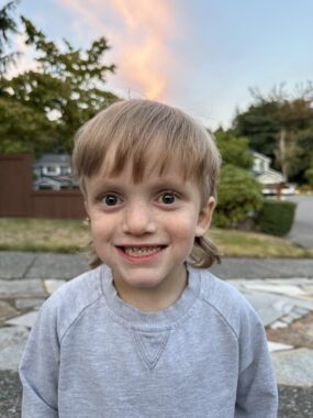 A young boy with blond hair and a gray sweatshirt puts on a smile as he stands outdoors in a neighborhood.