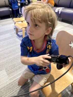 A young boy with a blue soccer jersey holds a video game controller in a hospital waiting room. He's looking up and to the right at a screen that's out of view. He's also excitedly half-sitting on a wooden chair and half-standing.