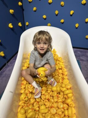 The young boy with blond hair and a gray sweatshirt sits in a bathtub filled with rubber ducks. Behind him is a dark blue wall that seems dotted with the yellow ducks.