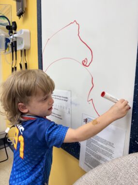 A 4-year-old boy draws with a red erase marker on a whiteboard in a doctor's office . 
