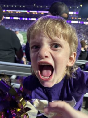 An extreme close-up of a young boy with his eyes and mouth wide open, cheering in excitement. Behind him is a purple-themed football stadium - the color of the home team. The boy is wearing a purple football jersey.