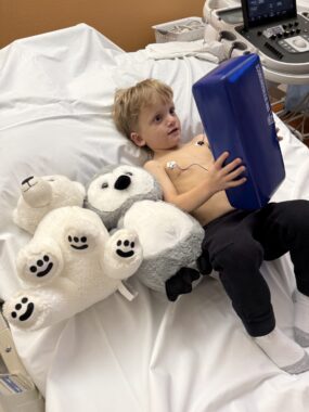 A young, skinny boy without a shirt lies on a hospital bed next to two large, white, stuffed animals. One is a polar bear and the other is a large owl. He's holding a big blue thing in his hands, but it's not clear what that is. He's clearly in a hospital setting but doesn't look to be stressed at all. 