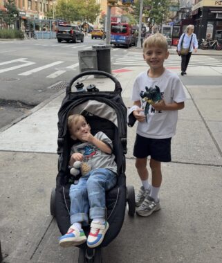 Two brothers, about 4 and 6 years old, pose for a photo on a New York City sidewalk. The older boy is standing and holding a toy, and the younger boy is sitting in a stroller and looking up at his big brother.