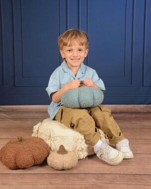A 4-year-old boy sits smiling broadly in a cute pre-K pose. His light blue shirt goes well with the dark blue background and the wooden floor. He's sitting on and holding stuff, soft, pillow-like pumpkins.