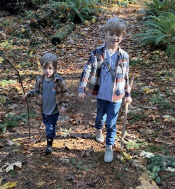 Two young boys walk on a leaf-lined trail in the woods. One is younger than the other and carries a long, skinny stick. Both are wearing matching flannel shirts. 