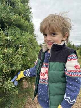A young boy smiles mischievously while standing next to a Christmas tree at what appears to be a tree farm. It looks chilly outside, as he's wearing a red and blue sweater with a green vest over it and gloves.