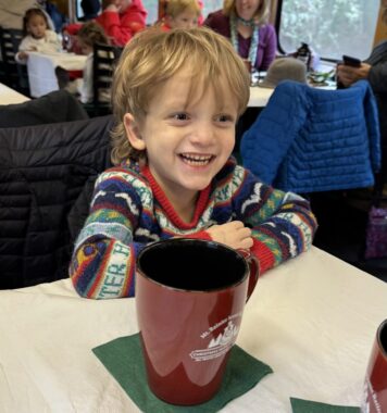 A young boy smiles broadly while looking just beyond the camera to the right. He's wearing a colorful holiday sweater and a large mug of hot chocolate sits in front of him. He's on a train, and several children and families are sitting in other seats behind him. 