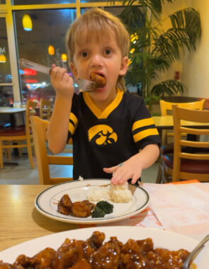 A young boy eats a bite of chicken at a table inside a restaurant. On the plate in front of him are a few more pieces of chicken, white rice, and broccoli.