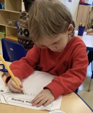 A 5-year-old boy sits at a table at day care, looking intently at something he's writing.