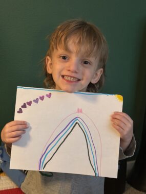 A boy smiles enthusiastically while holding up his colorful drawing, which includes a rainbow and several hearts. 