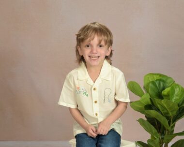A young boy sits for a photo in front of a neutral background. He has blond hair, is wearing a yellow button-down shirt, and is smiling happily.