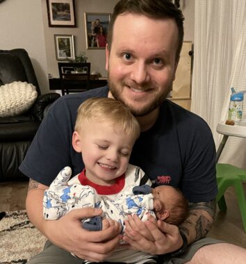 A man holds a young boy on his lap as the child holds an infant baby.