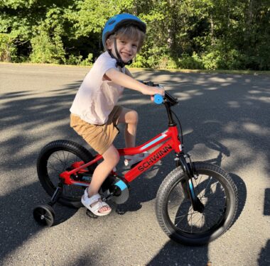 A young boy turns his head to smile as he pedals a bicycle with training wheels.