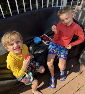 Two boys sit outside on a couch, eating strawberries. Both are wearing colorful swimming trunks, long-sleeved shirts, and sandals. 