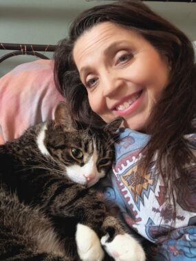 A woman reclines on her bed and takes a selfie of herself and her cat, who is snuggled up next to her.