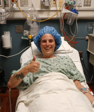 A woman lies on a hospital bed and gives the thumbs-up sign.