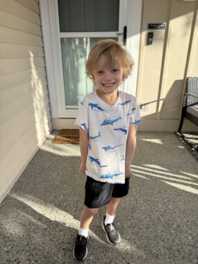 A 5-year-old boy smiles and shows off his shirt, which is white and features blue sharks. He looks to be standing outside the front door of his home.