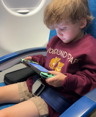 A child sits at the window seat of an airplane, playing on his e-tablet.