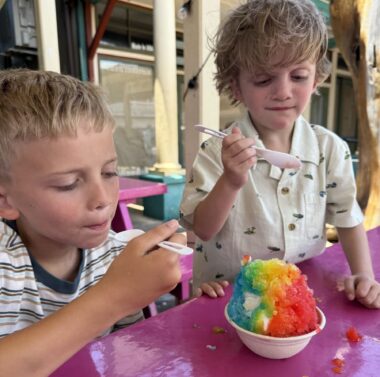 Two young boys sit at an outdoor table eating rainbow-colored shaved ice.