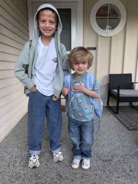 Two brothers, about 8 and 5 years old, stand side by side in front of their home on the first day of school. Both have on their backpacks and are smiling excitedly.