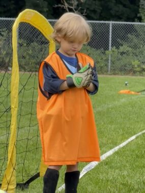 A young boy in a sports team uniform stands in front of a goalie net. The orange jersey hangs down to his knees, and he's looking down at the gloves he's wearing.